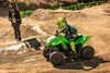 Side angle of a person riding an ATV on a closed dirt course with parental supervision in the background.