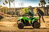 Side angle of a person standing while riding an ATV on a closed dirt course with parental supervision in the background.