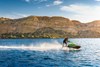 Side angle of a person on a personal watercraft on the water in front of a mountain.