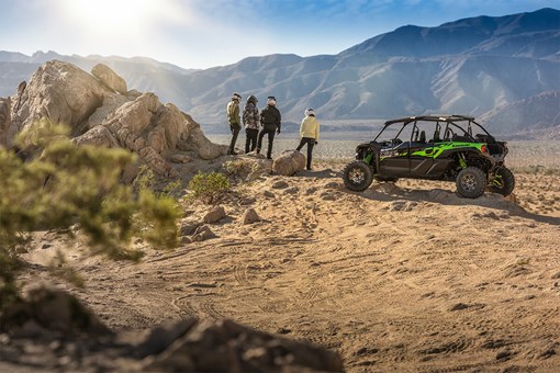 Three-quarter rear angle of four people standing next to a side x side off-road. opens in a new window