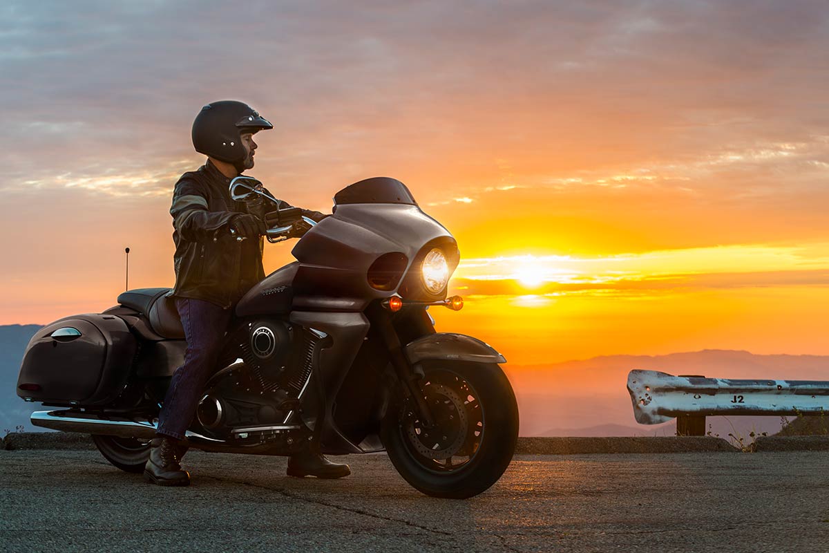 Three-quarter front angle of a person sitting on a parked motorcycle in front of a sunset.