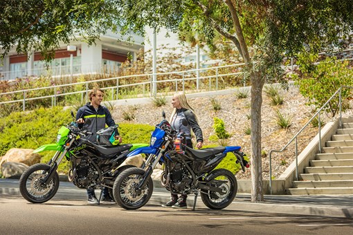 Three-quarter front angle of two people standing next to their motorcycles in a parking lot. opens in a new window