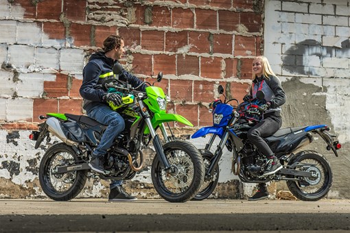 Three-quarter front angle of two people sitting on parked motorcycles in an urban city. opens in a new window