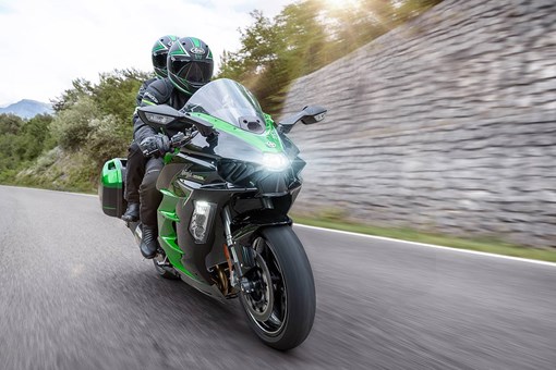 Three-quarter front angle of two people riding on a motorcycle on a highway next to a stone wall. opens in a new window