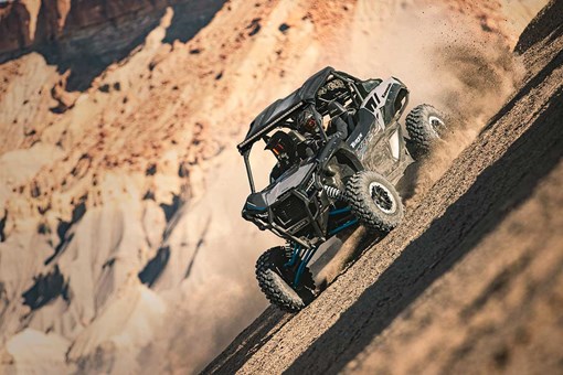 Three-quarter front angle of a person and passenger driving a side x side with blue accents off-road. opens in a new window