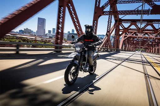 Three-quarter front angle of a person riding a motorcycle across a city bridge. opens in a new window