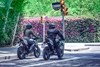 Three-quarter rear angle of two people on motorcycles stopped at a red light.