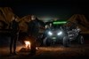 A group of people talking around a bonfire at night with a side x side parked in the background.