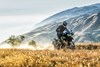 Three-quarter front angle of a person riding a motorcycle on a dirt road with mountains in the background.