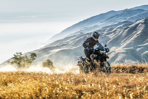 Three-quarter front angle of a person riding a motorcycle on a dirt road with mountains in the background. opens in a new window