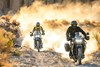 Front angle of two motorcycles riding off-road with dust in the background.