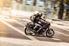 Side angle of a person and a passenger riding a motorcycle on a city street.