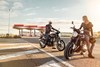 Front angle of two riders with parked motorcycles near a gas station.