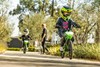 Three-quarter front angle of two kids riding electric balance bikes down a neighborhood street with a parent supervising.