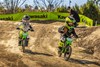 Three-quarter front angle of two kids riding a electric balance bikes on a dirt track with an adult supervising.