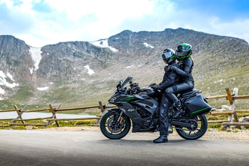 Side angle of a rider and passenger seated on a parked motorcycle. opens in a new window
