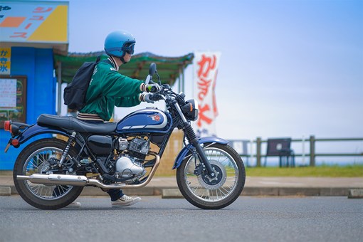 Side angle of a person standing near a blue motorcycle. opens in a new window