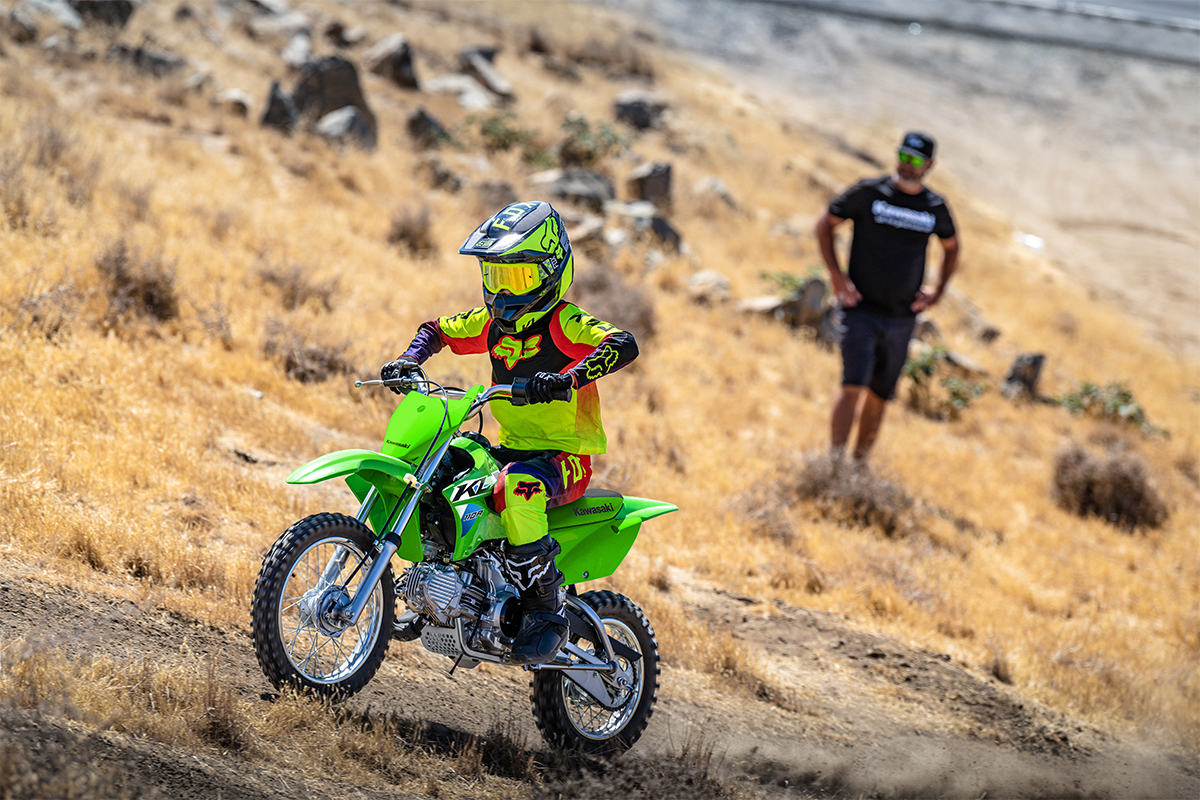 Side angle of a person riding a green motorcycle off-road under adult supervision.