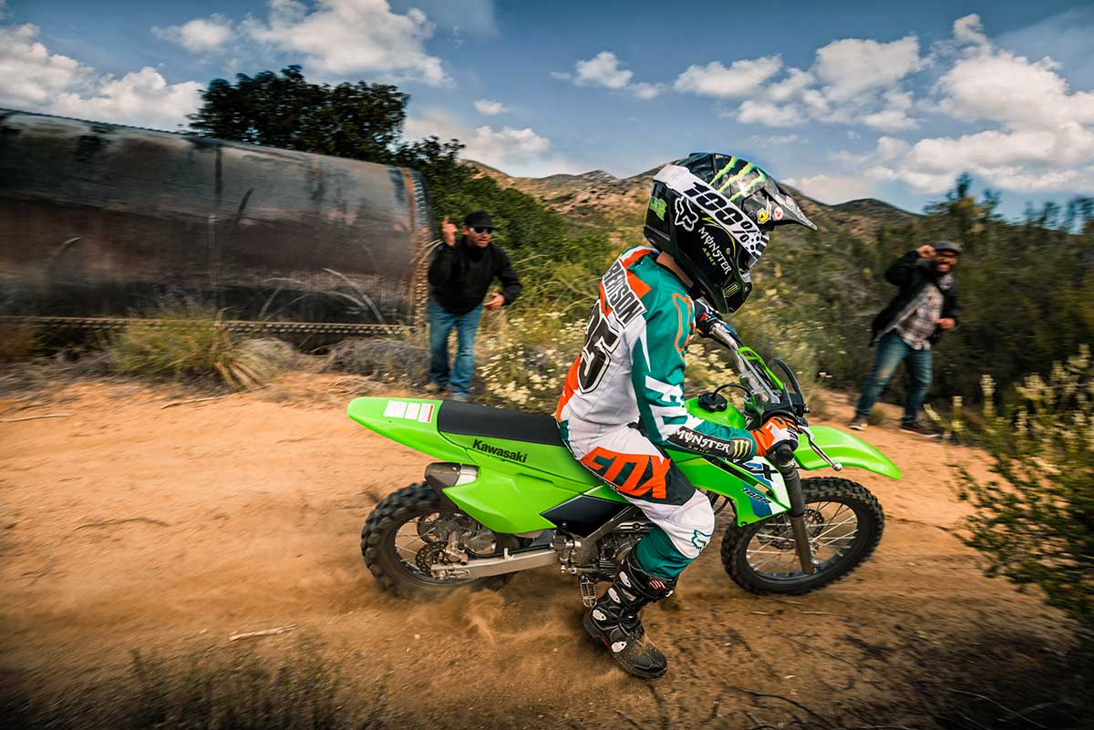 Side angle of a person riding a green motorcycle off-road under adult supervision.