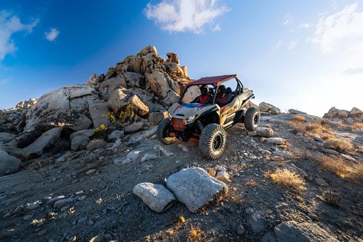 Three-quarter front angle of two people riding down an off-road decline in a side x side. opens in a new window