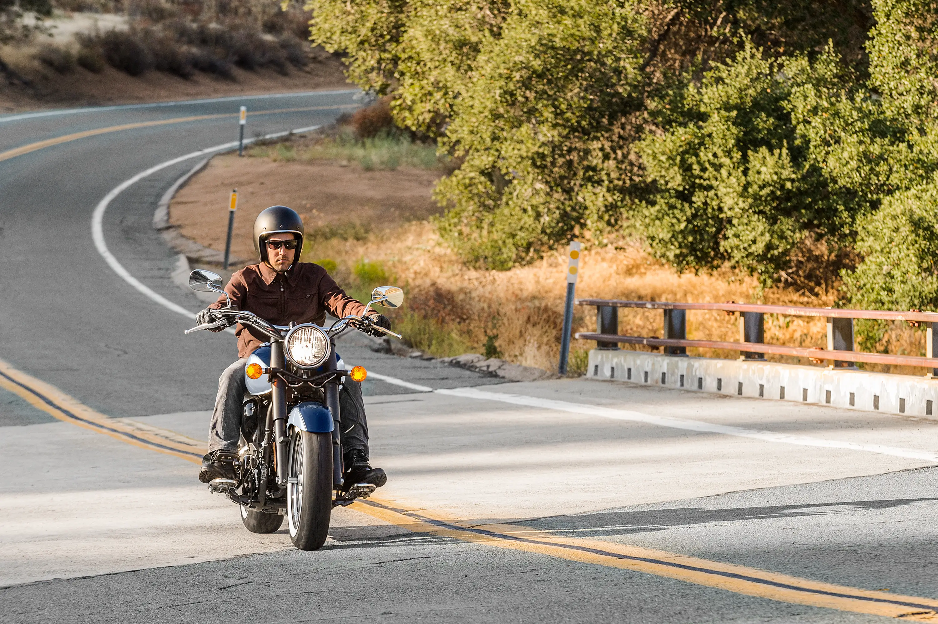 Front three-quarter angle of a person riding a motorcycle over a bridge.