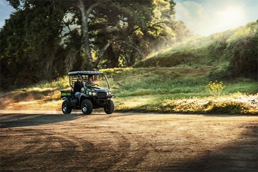 Two people riding in a side x side across a dirt lot with a tree in the background. opens in a new window