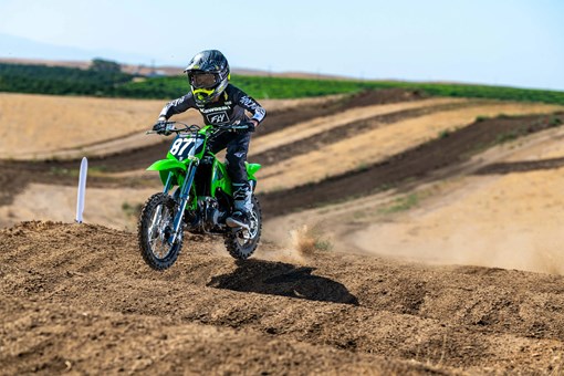 Front three-quarter left view of a kid riding a green youth motocross dirt bike on a track over bumps. opens in a new window