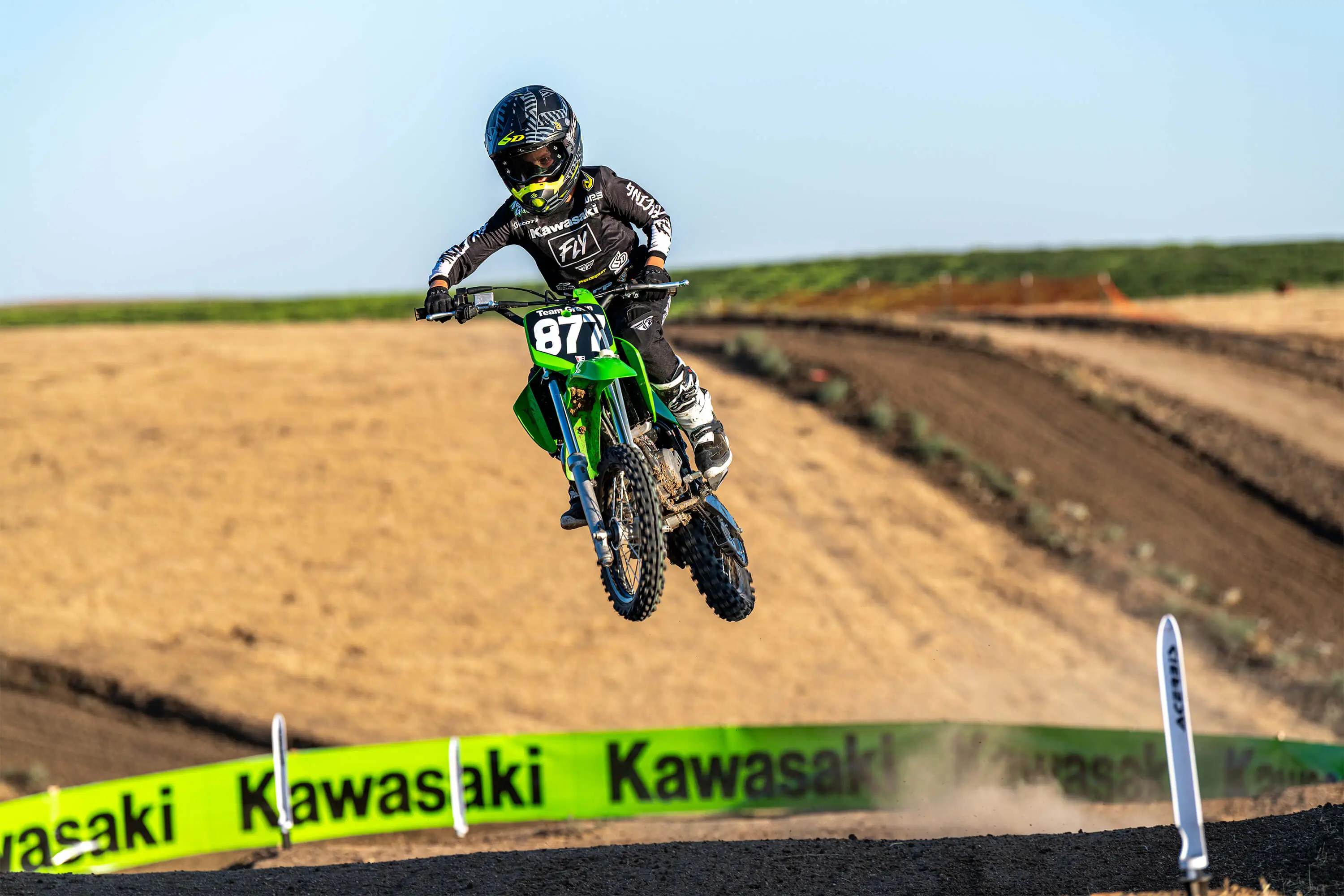 Oncoming view of a kid riding a green youth dirt bike over a jump on a motocross track.