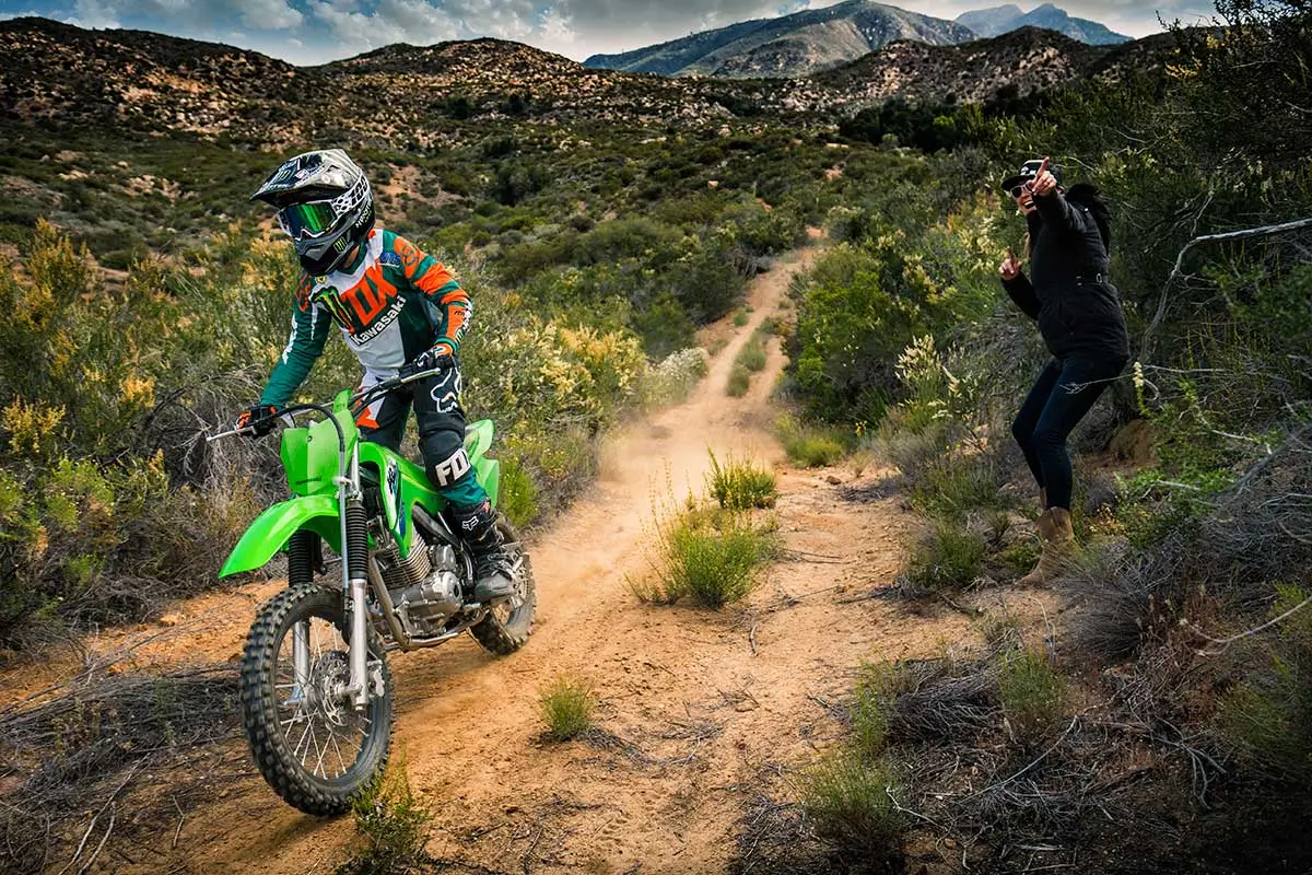 A kid riding a green dirt bike on a trail with a parent watching in the background.