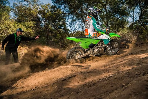 Side angle of a kid riding a dirt bike up a hill with a parent watching in the background. opens in a new window