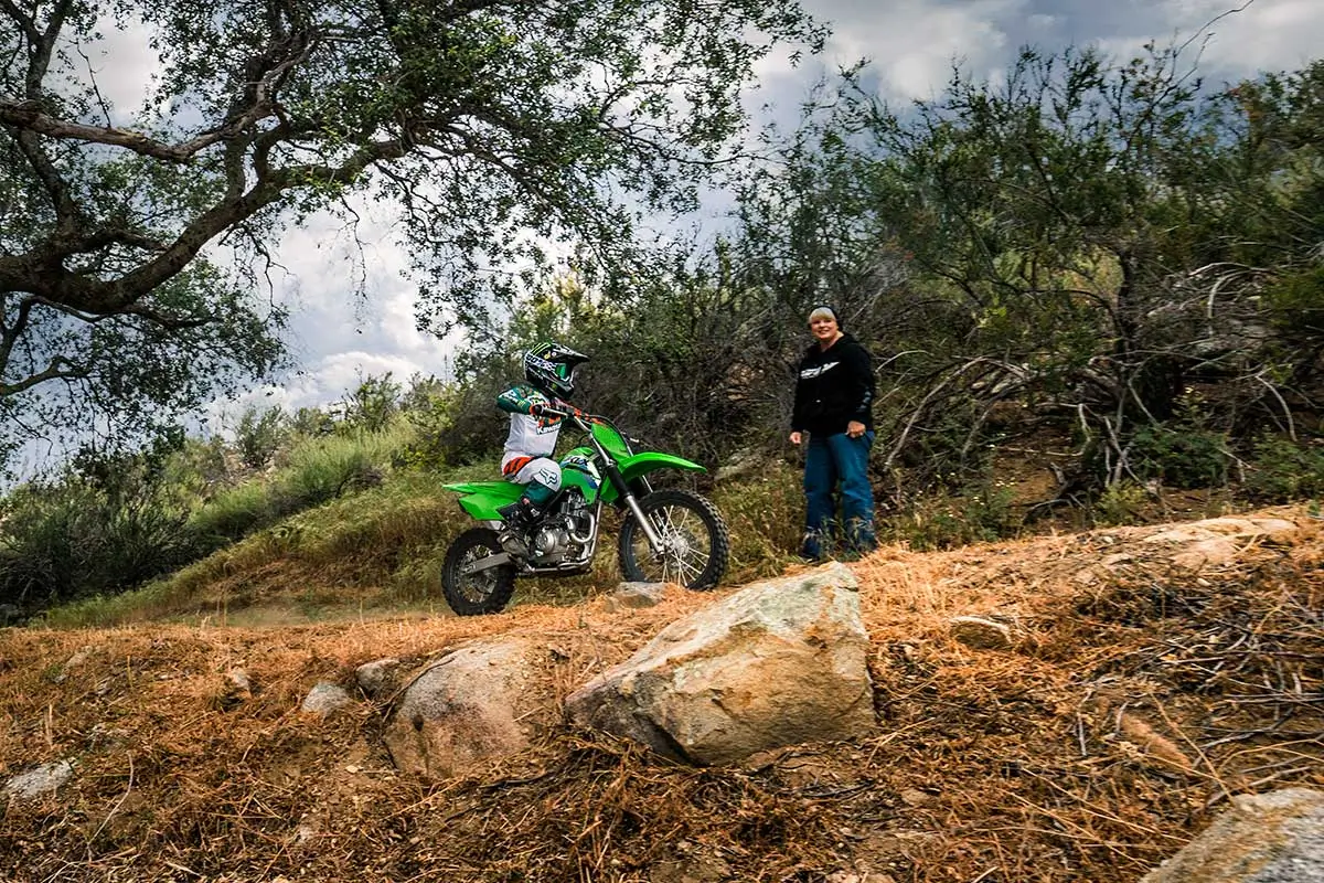 A kid a green dirt bike riding on a trail next to a parent.