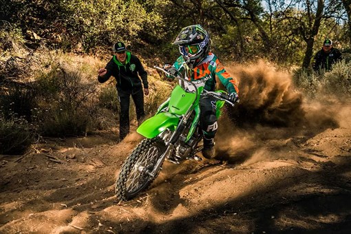 Oncoming view of a kid riding a dirt bike off-road with a parent watching in the background. opens in a new window