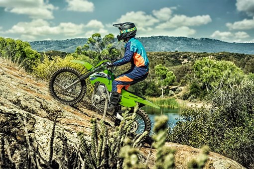 Side view of a person riding a green dirt bike up a hill in a forest. opens in a new window