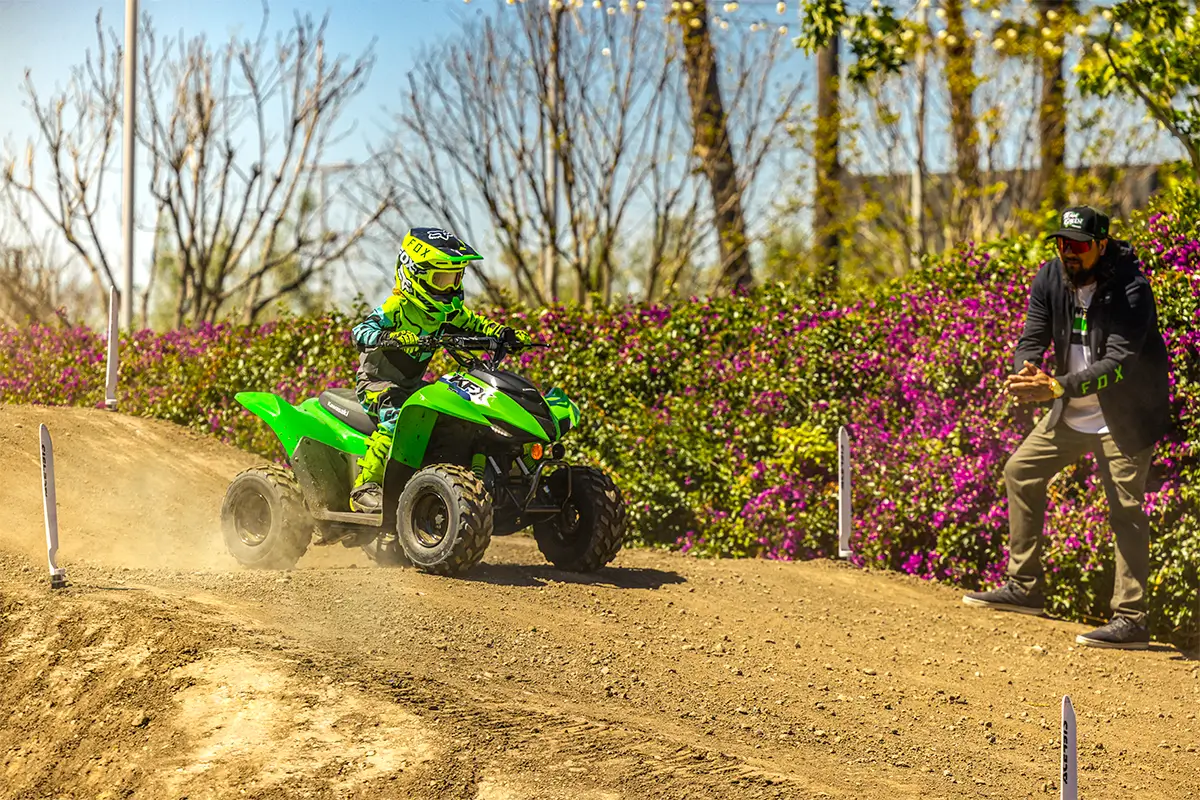 A kid riding a green youth ATV down a hill on a track as a parent gives encouragement.