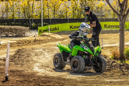 A kid riding a green youth ATV through turns on a track in front of a parent. opens in a new window