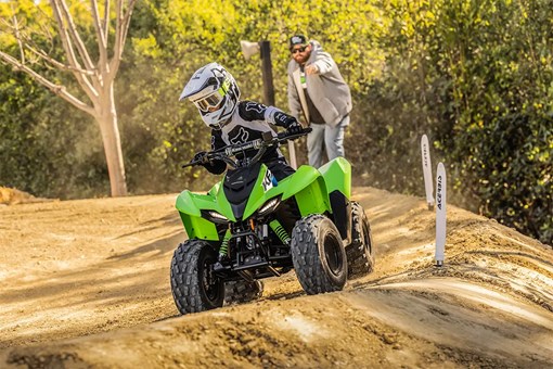 A kid riding a green youth ATV through a turn on a track as a parent gives instruction. opens in a new window