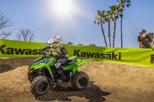 A kid riding a green youth ATV on a motocross track with a parent in the background. opens in a new window