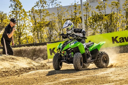 A kid riding a green youth ATV around a turn on a track with a parent in the background. opens in a new window