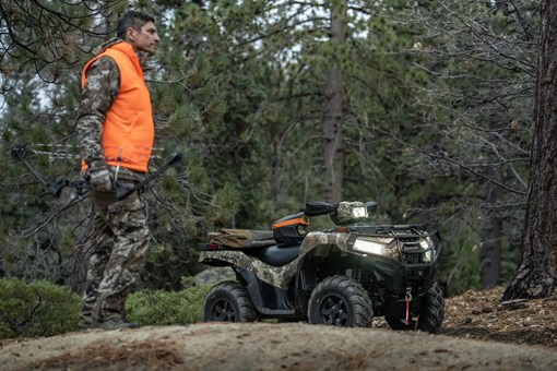 Person wearing camo and an orange vest in front of a utility ATV parked in the woods. opens in a new window