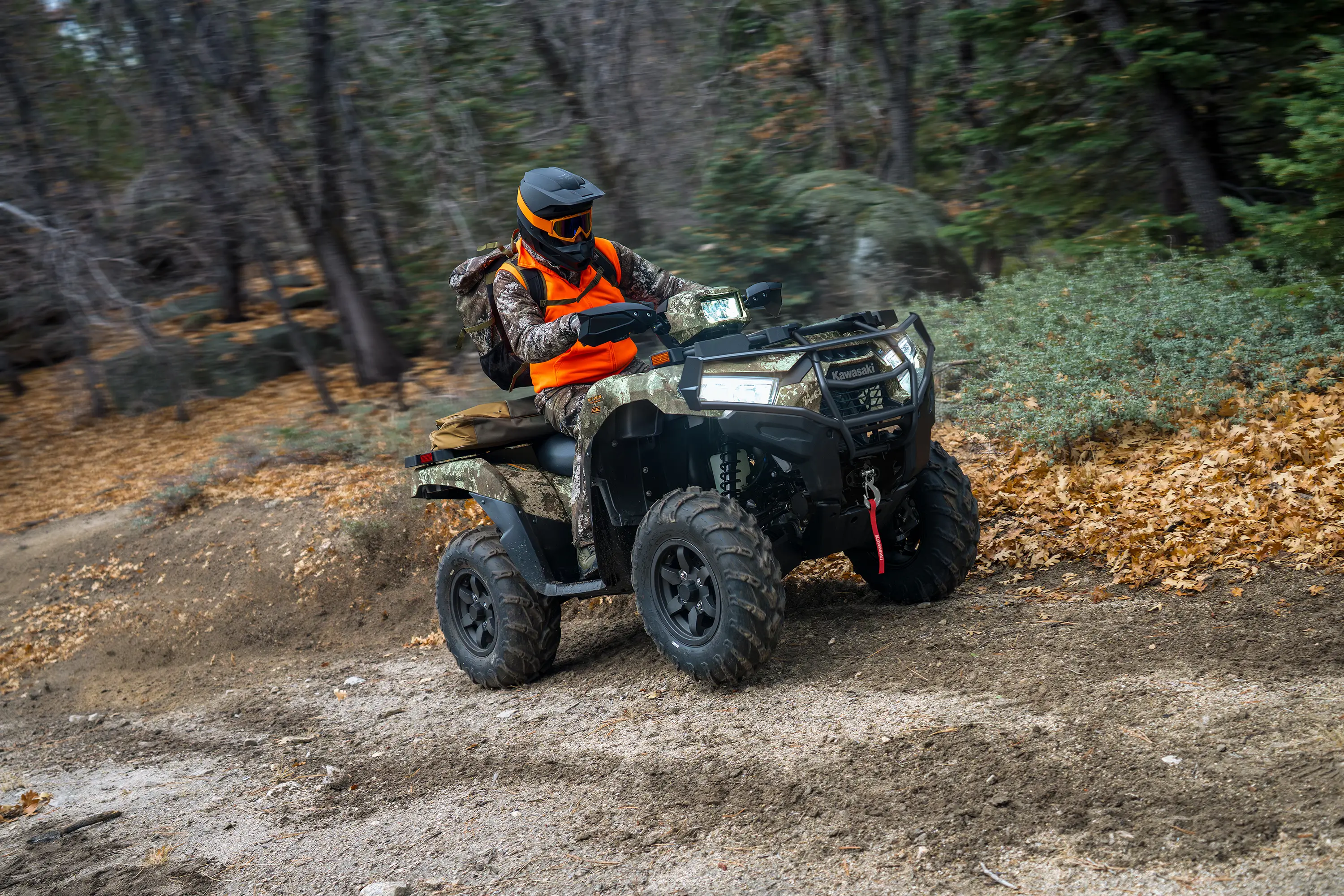 Right side view of a person wearing an orange vest riding a utility ATV on a road through the woods.