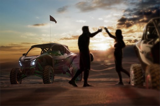 Three-quarter rear angle of two people and a side x side parked on sand. opens in a new window
